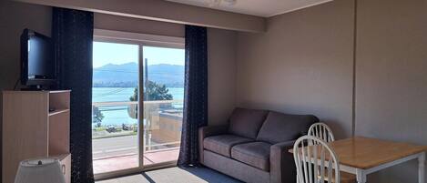A room with a sofa, a table, a chair, a ceiling fan, a TV on a cabinet, and a view of a body of water and mountains.