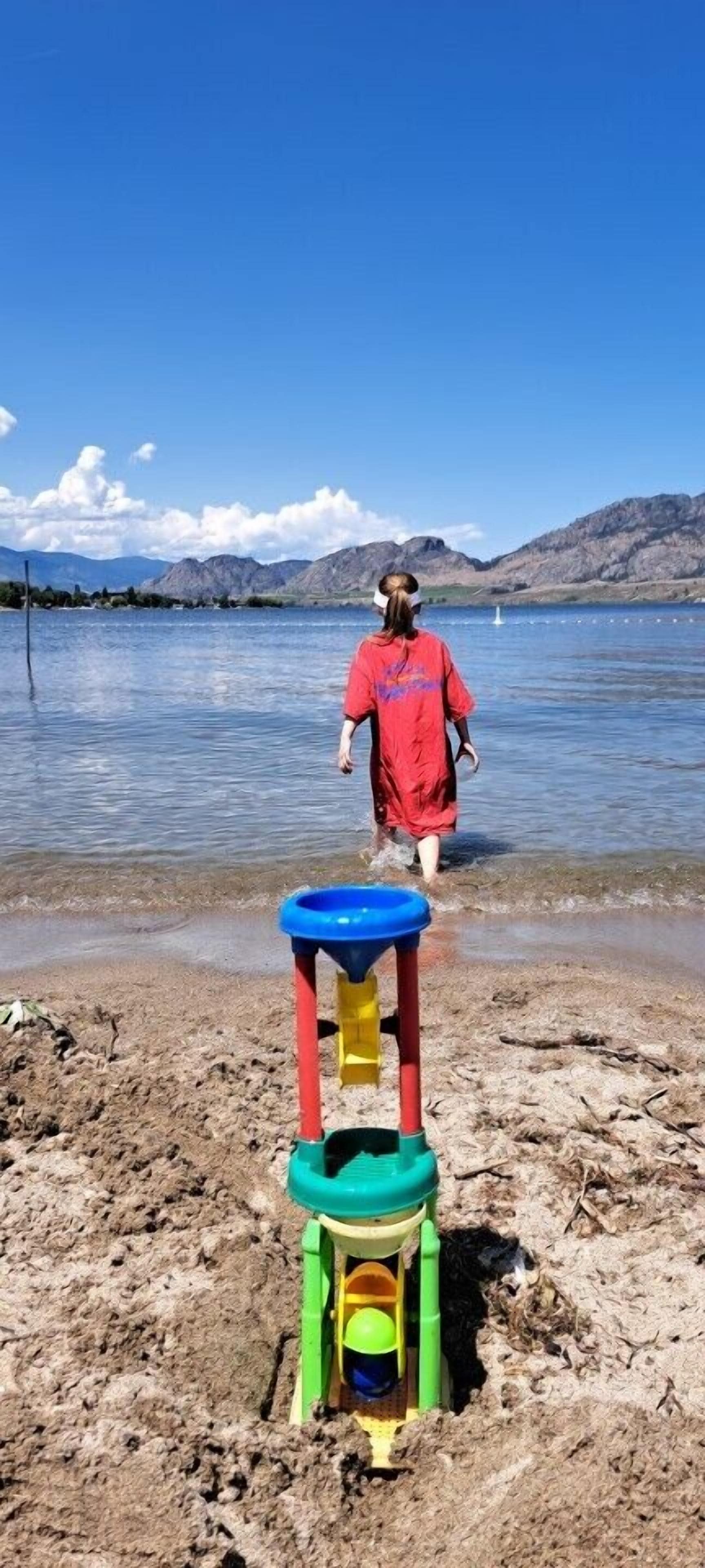Niño con camisa roja caminando hacia una playa con un castillo de arena y agua de fondo.