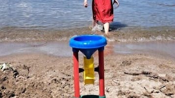 Niño con camisa roja caminando hacia una playa con un castillo de arena y agua de fondo.
