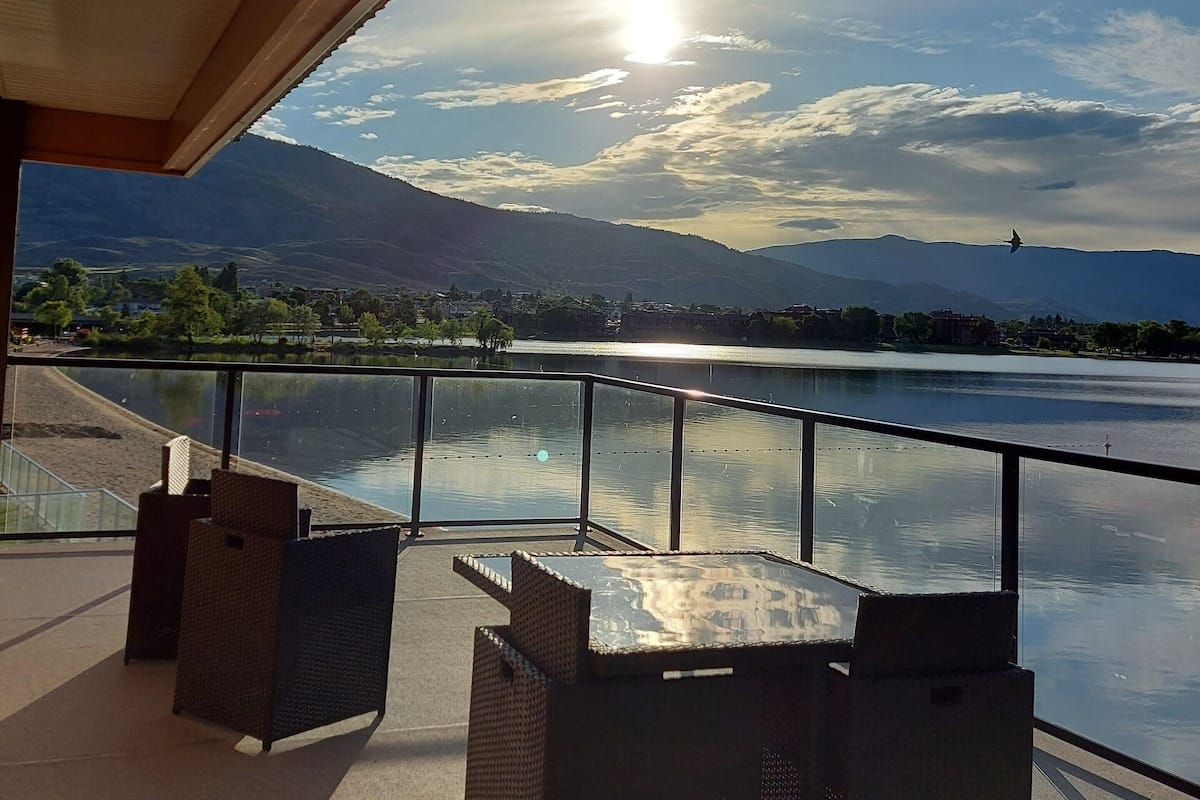 A terrace with wicker furniture overlooking a lake, with mountains in the background and a clear sky.