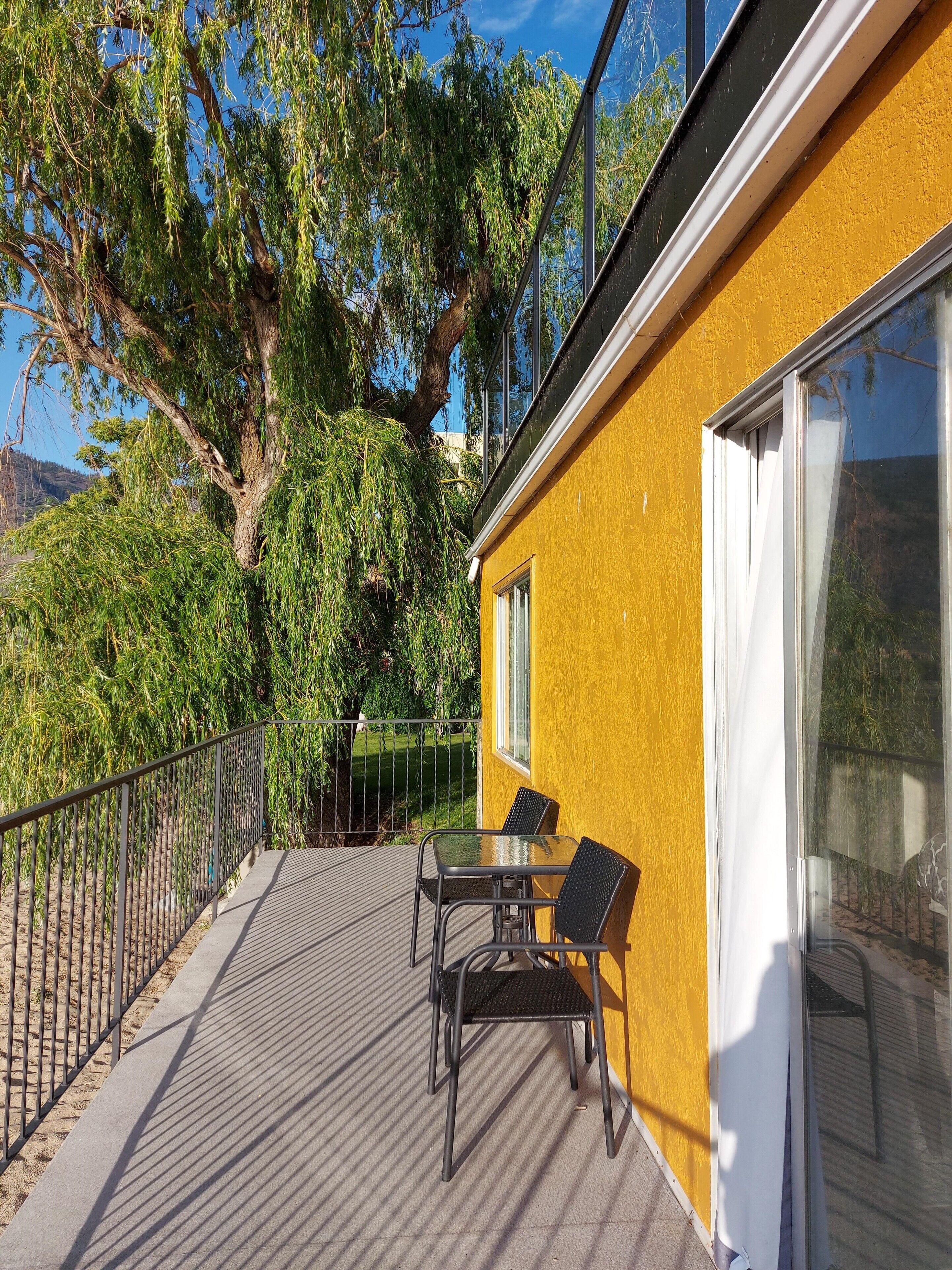 A balcony with a table and chairs, a yellow wall, and a large tree in the background.