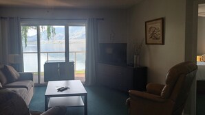 Een woonkamer met een bank, fauteuil, tv-meubel, een tafel en uitzicht op een meer door grote ramen.