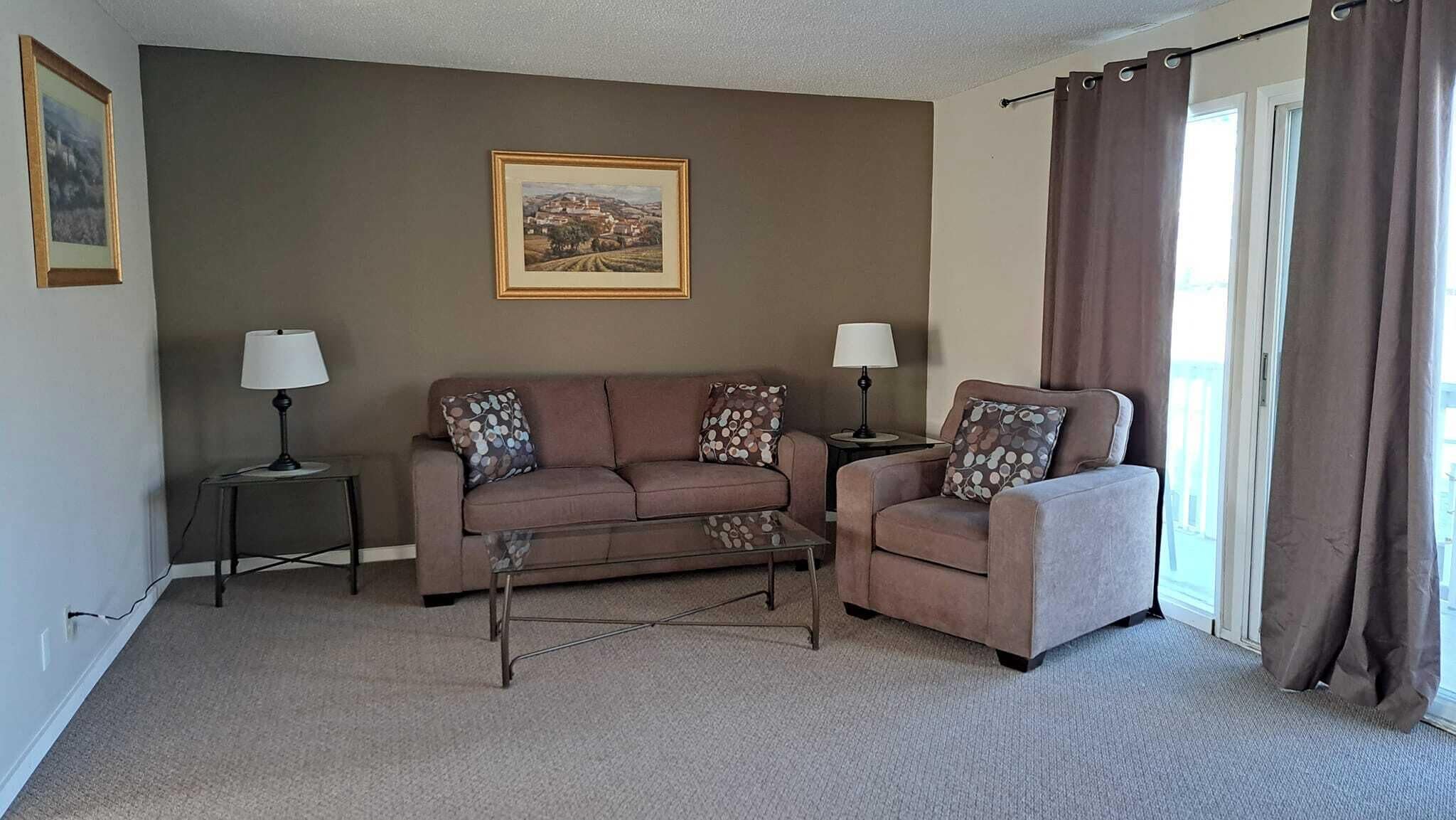 A living room with a brown sofa, a smaller armchair, two side tables with lamps, a coffee table, and a framed picture on the wall.