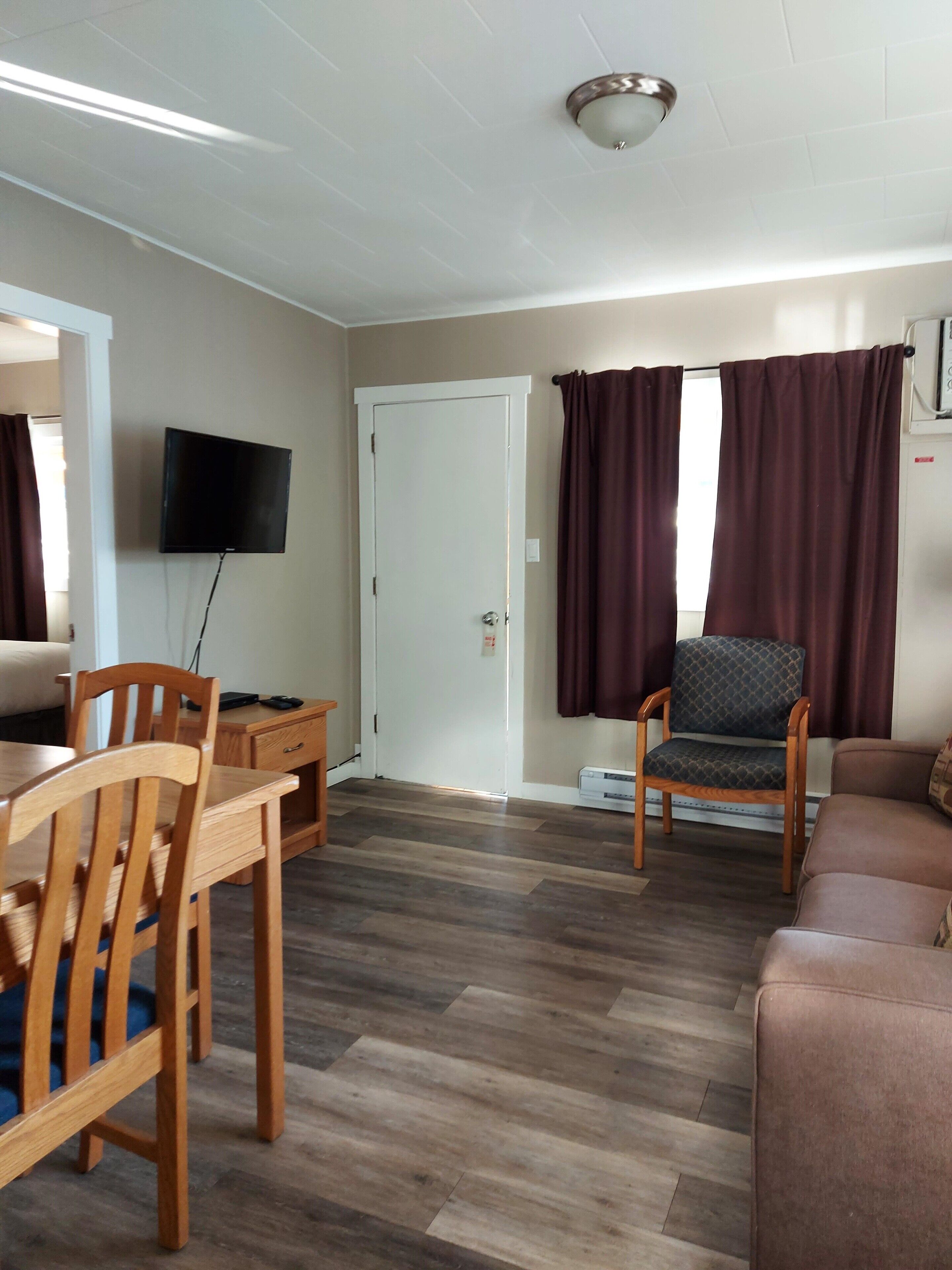 A living room with a wooden dining table, chairs, a sofa, a TV mounted on the wall, and a door with a window.