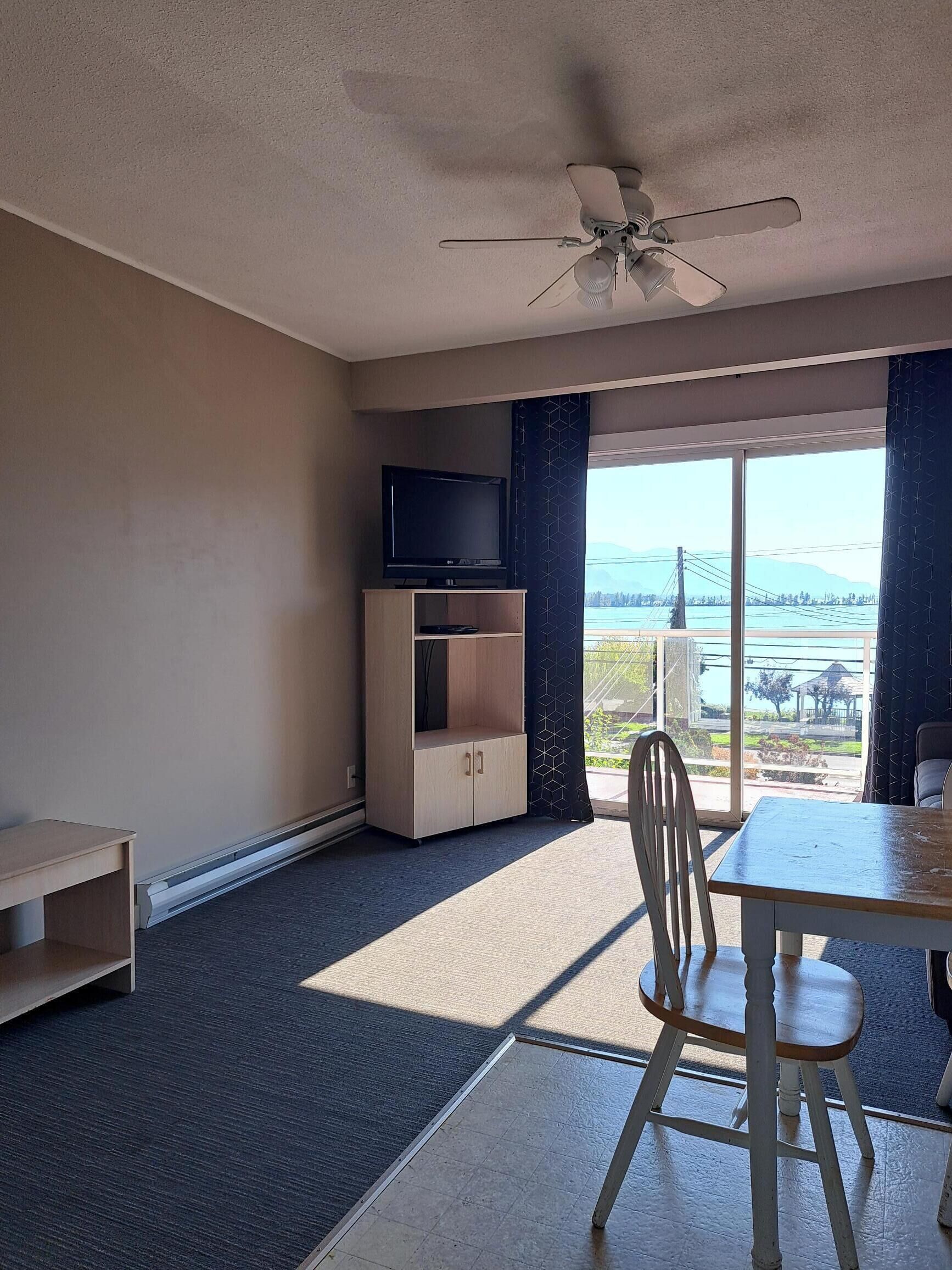 A hotel room with a large window, a ceiling fan, a TV on a wooden cabinet, and a dining table with chairs.