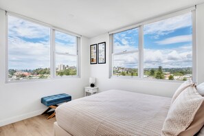 Ein Schlafzimmer mit einem großen Bett, einem kleinen Nachttisch, einer blauen Bank und zwei großen Fenstern mit Blick auf die Stadt.