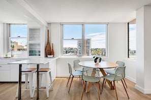 Une cuisine moderne avec des meubles blancs, un coin repas doté d’une table ronde et de chaises en fil de fer, et de grandes fenêtres offrant une vue sur la ville.