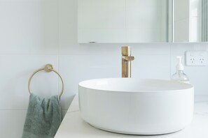Ein helles Badezimmer mit einem runden Waschbecken, einem goldenen Wasserhahn, einem Handtuchhalter mit einem grünen Handtuch und einem Spiegel über dem Waschbecken.