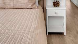 Un dormitorio ordenado con cama, almohadas, una mesita de noche con lámpara y un mueble auxiliar.