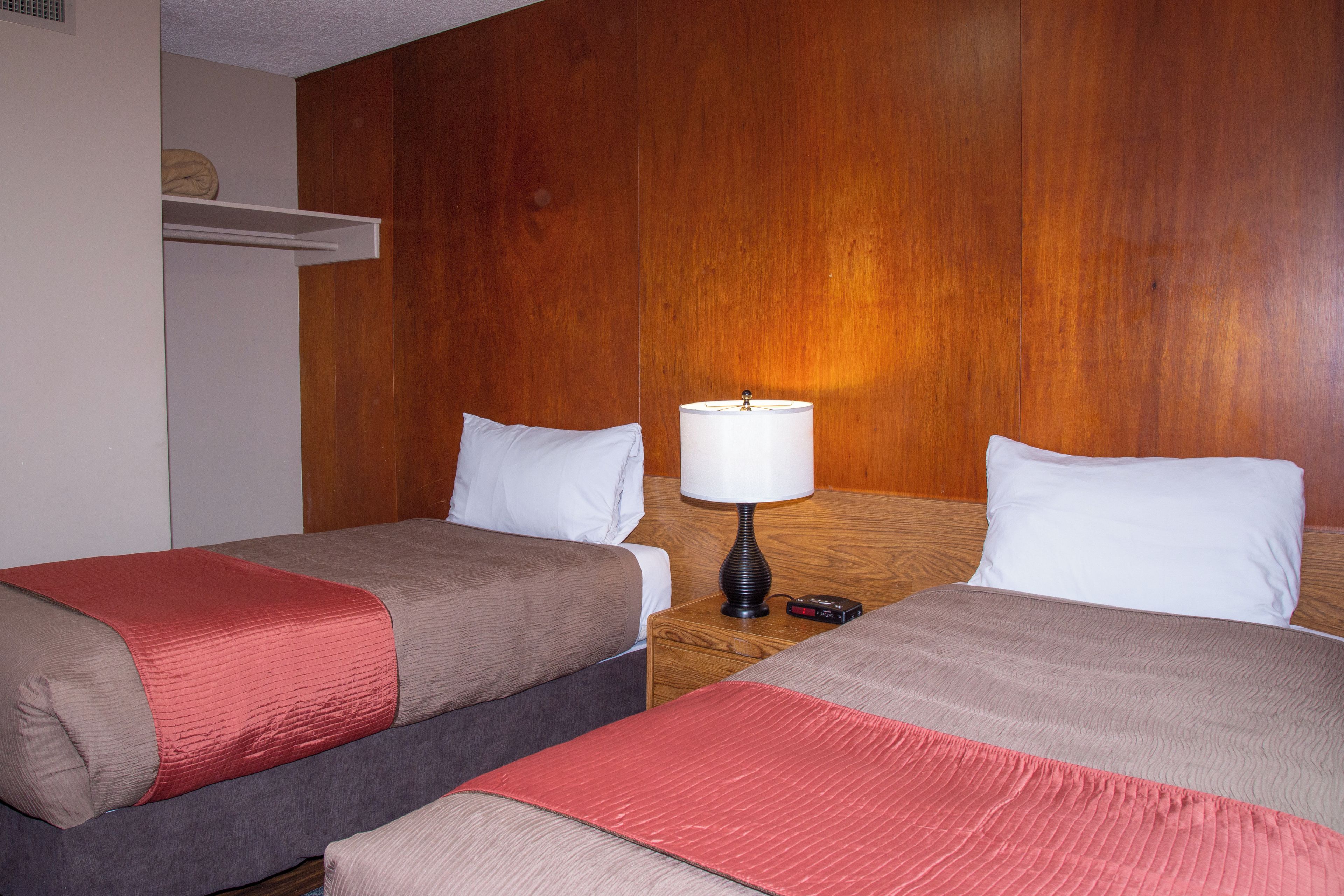 Habitación de hotel con dos camas, cada una con una almohada y una manta. Un mesita de noche con una lámpara y un reloj se encuentra entre las camas.