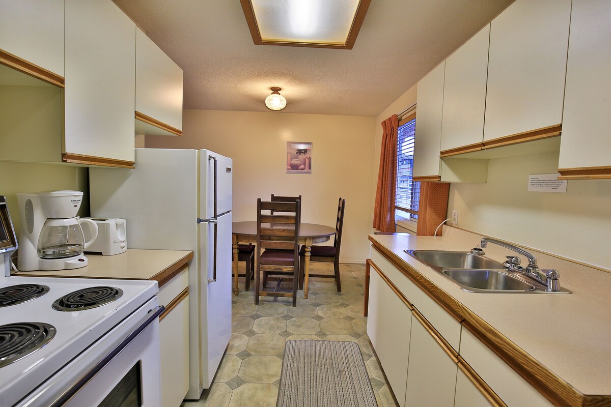 A compact kitchen with a stove, refrigerator, and sink. There is a dining table with chairs and a coffee maker.