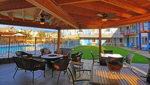 Un patio al aire libre con mesas y sillas de madera, una piscina y vistas a un edificio de colores azul y blanco.