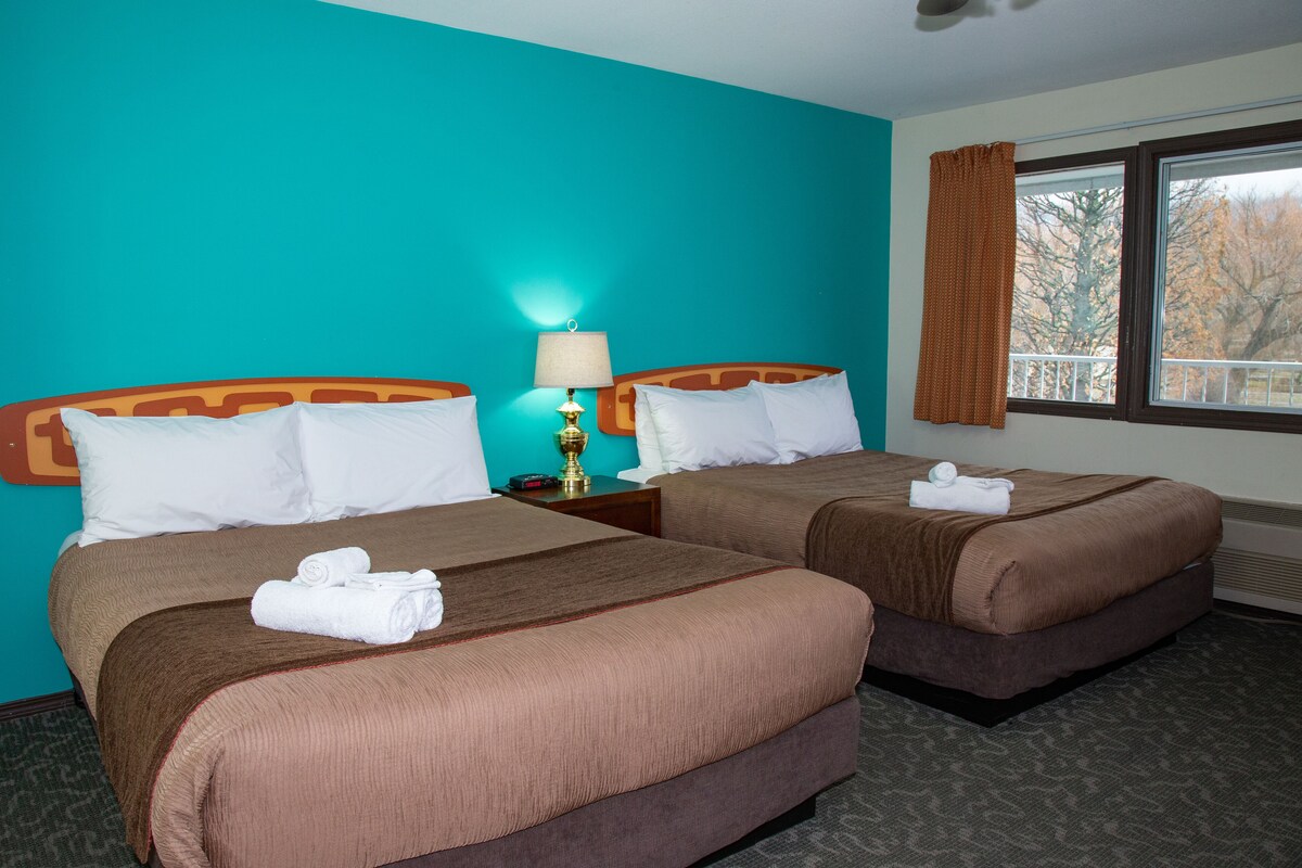 A hotel room with two beds, teal walls, and a window with a view of trees.