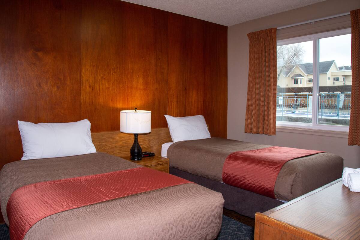 A hotel room with two beds, a nightstand with a lamp, a window with curtains, and a view of houses outside.