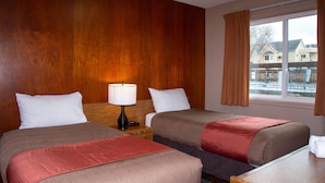 Habitación de hotel con dos camas, una mesita de noche con lámpara, una ventana con cortinas y vistas a casas.