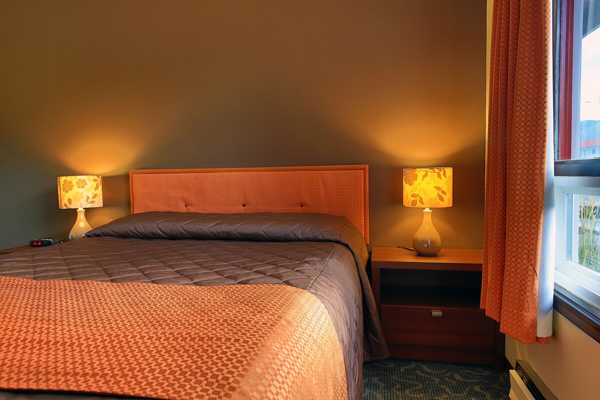 Una habitación de hotel con una cama, dos lámparas de noche, una mesita de noche y una ventana con cortinas.