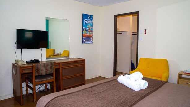 A hotel room with a bed, a desk with a chair, a television, and a door leading to a bathroom. - Bowmont Motel (Penticton)
