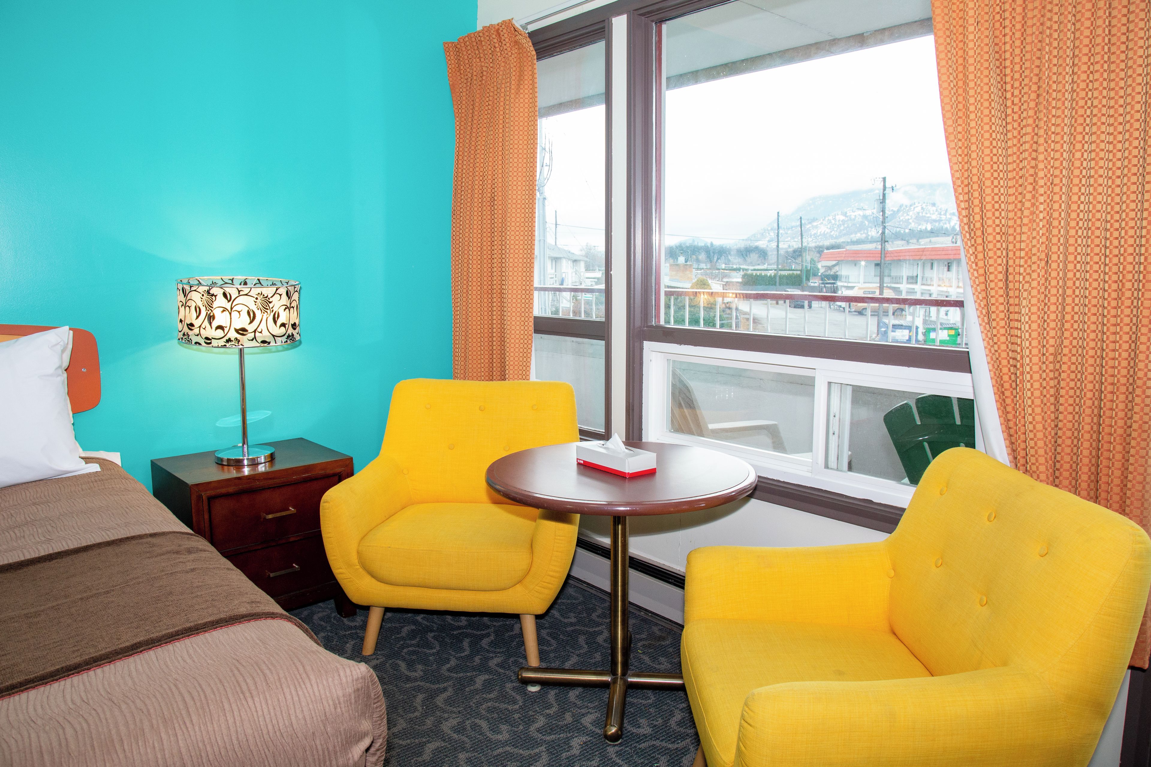 Una habitación de hotel con una cama, dos sillones amarillos, una mesita, una lámpara y un ventanal con cortinas.