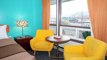 Una habitación de hotel con una cama, dos sillones amarillos, una mesita, una lámpara y un ventanal con cortinas.