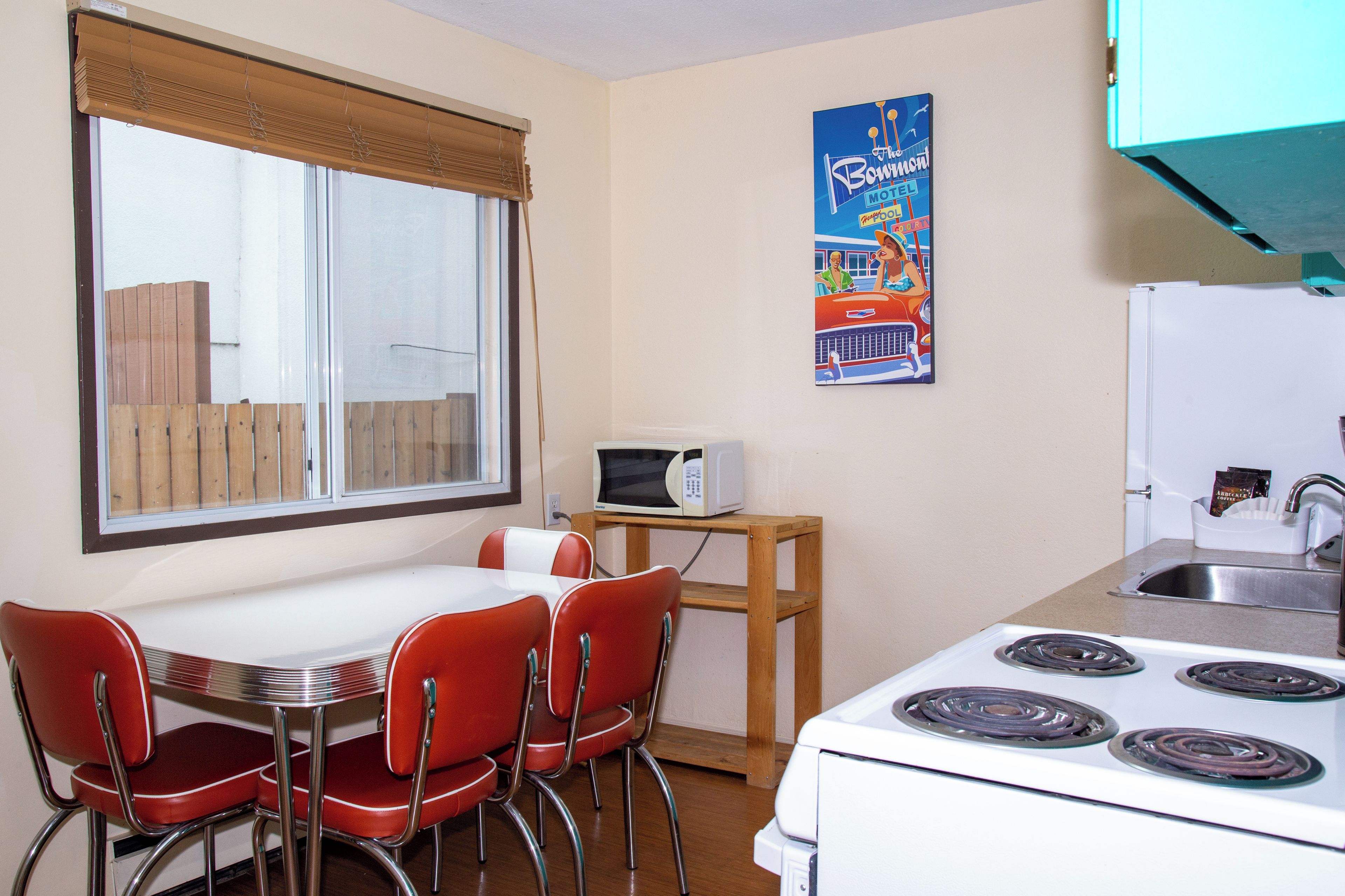 A compact kitchen with a white stove, red chairs, a microwave on a wooden shelf, and a window with blinds.