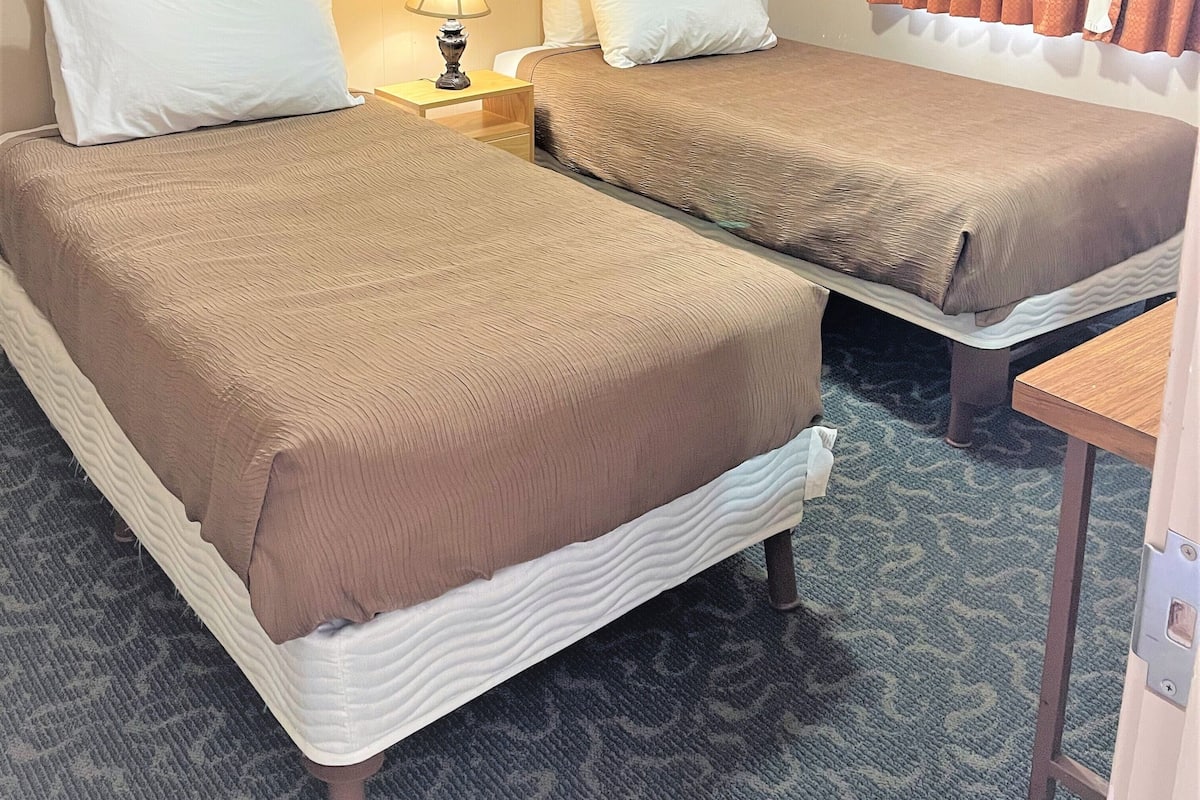A hotel room with two beds, a nightstand with a lamp, a small table, and a window with curtains.