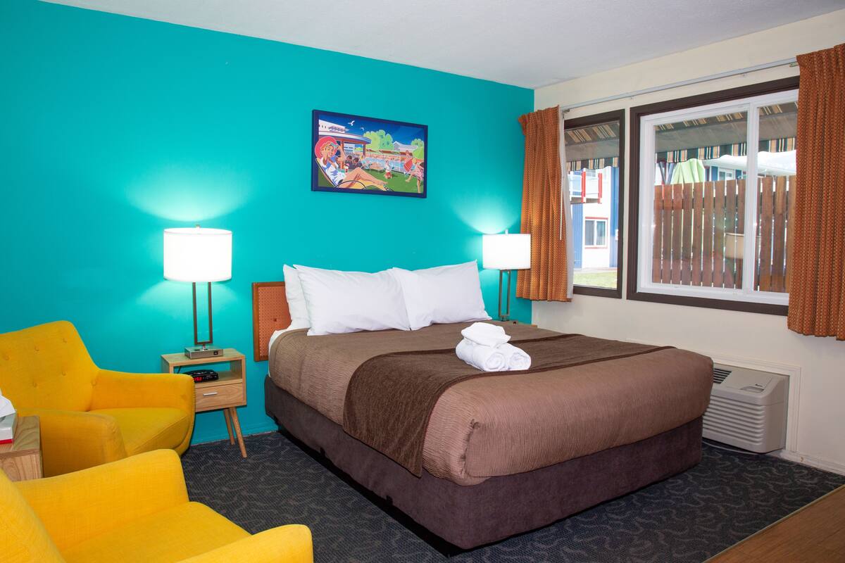 A hotel room with a large bed, two bedside lamps, a bright yellow armchair, and a window with curtains.