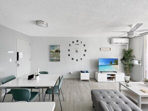 Een moderne woonkamer met een eethoek, een grote flatscreen-tv, een plafondventilator en een wandklok.
