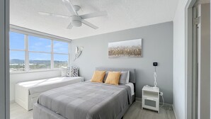 A modern bedroom with a large bed, a bedside table, a ceiling fan, and a view of the outdoors. - Exceptional Modern Beachfront Escape • XL Pool • In-Unit Laundry • VIP Parking (Carolina)