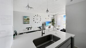 A modern kitchen with a white countertop, black sink, and pendant lights. There is a clock on the wall and a painting above the counter. - Exceptional Modern Beachfront Escape • XL Pool • In-Unit Laundry • VIP Parking (Carolina)