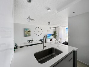 Una cocina moderna con encimera blanca, pileta negra y lámparas colgantes. Hay un reloj en la pared y un cuadro sobre la encimera.