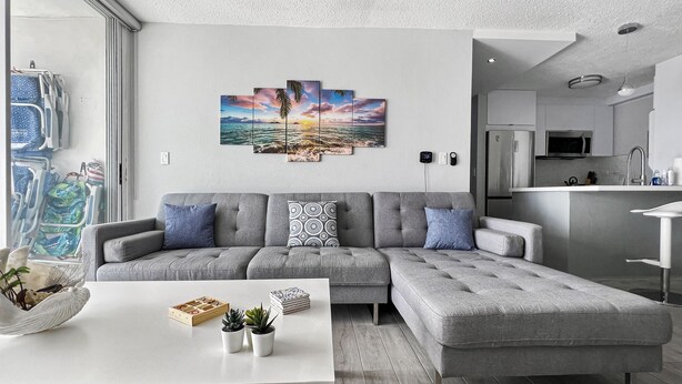 A modern living room with a grey sectional sofa, a white coffee table, and a wall-mounted artwork depicting a sunset over the ocean. - Exceptional Modern Beachfront Escape • XL Pool • In-Unit Laundry • VIP Parking (Carolina)