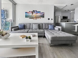 Een moderne woonkamer met een grijze modulaire bank, een witte salontafel en een kunstwerk aan de muur met daarop een zonsondergang boven de oceaan.