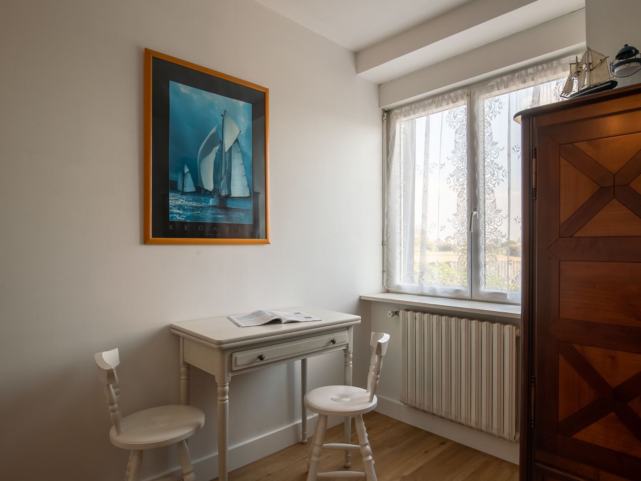 Una habitación con un escritorio blanco, dos sillas blancas, un radiador y una ventana con cortinas de encaje.