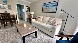 Ein Wohnzimmer mit einem beigen Sofa, einem Couchtisch, einem blauen Sessel, einer Lampe und einem Gemälde mit Strandmotiv.