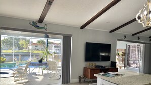 A spacious living room with a large TV, a dining area with chairs and a table, a kitchen with a dishwasher, and a view of a swimming pool and greenery outside. - Punta Gorda Isles Jewel with 45ft dock,just4minutes from Fishville and downtown. (Punta Gorda)