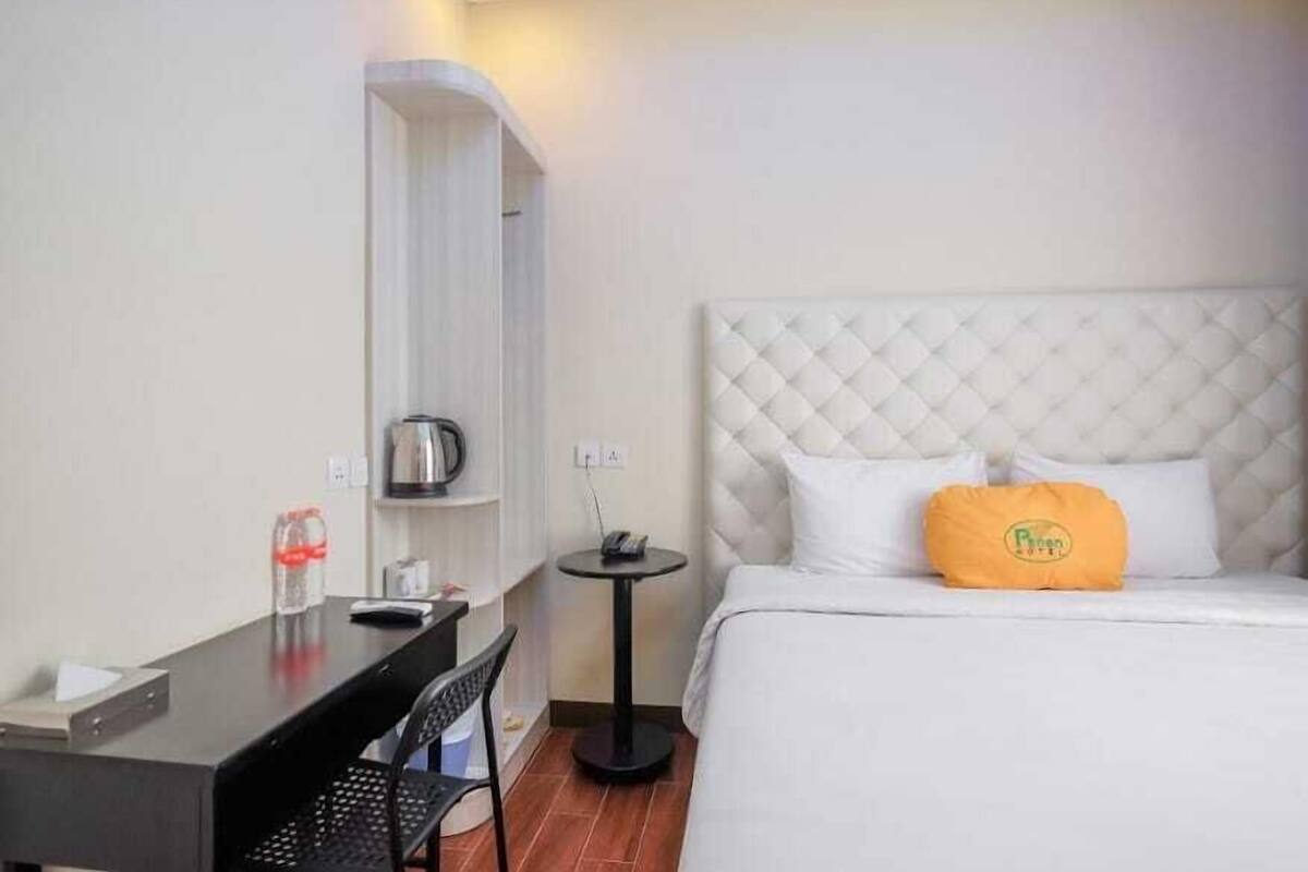 A hotel room with a bed, a small table, a chair, a shelf, and a kettle.