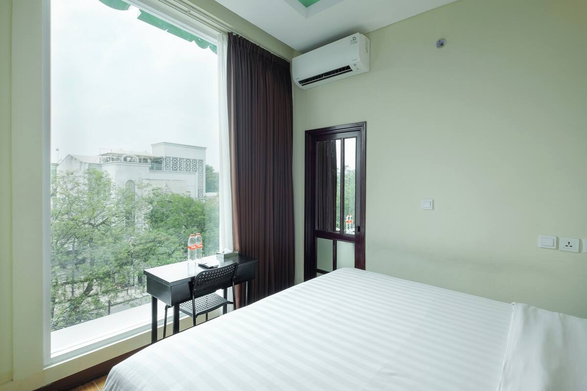 A hotel room with a bed, a desk with a chair, a window with curtains, and an air conditioning unit.