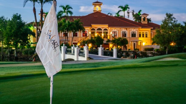 Un terrain de golf avec un drapeau, des palmiers et un grand bâtiment à l’arrière-plan, dont le toit est rouge.