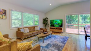 A modern living room with a sofa, coffee table, and TV. There are large windows with blinds, a plant, and a view of a deck. - Large 5BR House w/ Backyard, Deck, and Patio (Princeton)
