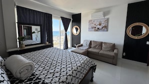 A bedroom with a bed, a sofa, a large window with curtains, a mirror, and a wall-mounted air conditioner.