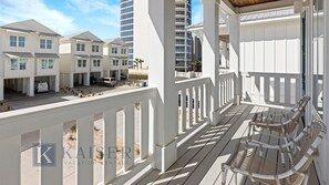 Une propriété située en bord de mer, avec des bâtiments en bois peints en blanc, un balcon couvert agrémenté de meubles en osier et une vue dégagée sur l’océan.