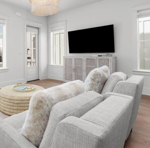 Un salon moderne avec un grand canapé d’angle gris, un téléviseur à écran plat fixé au mur et un pouf tressé surmonté d’un plateau.