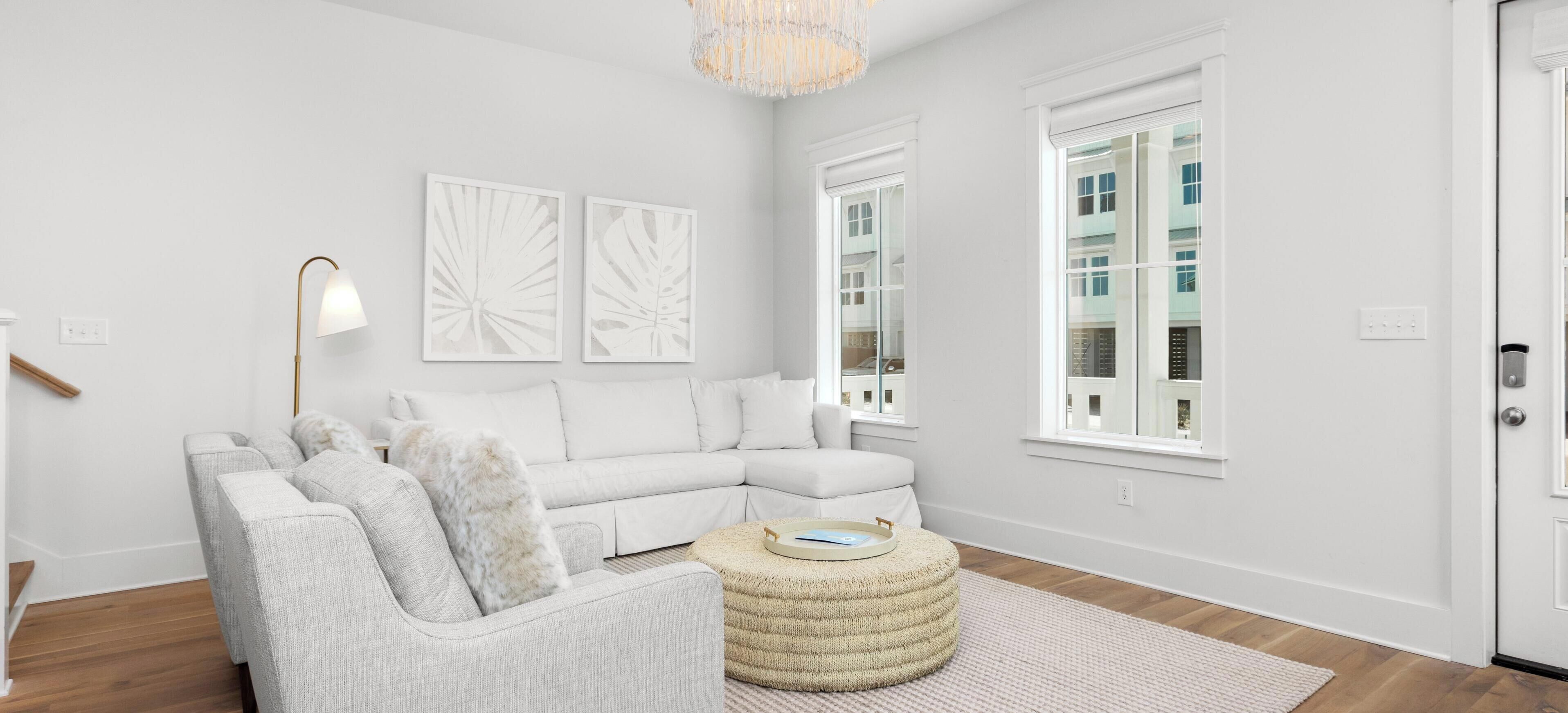 A modern living room with a grey sectional sofa, a round ottoman, and a large rug. There are two windows with white frames, and a chandelier hangs from the ceiling.