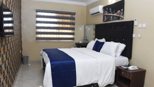 A hotel room with a bed, a nightstand, a wall-mounted air conditioner, a window with blinds, and a cityscape painting. - Captivating 1-bed Apartment in Awoyaya (Lekki)