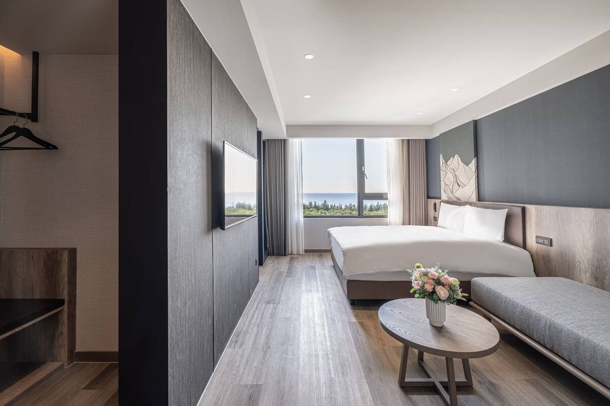 Quarto de hotel moderno com uma cama grande, um sofá, uma mesinha com um vaso e vista para o mar.