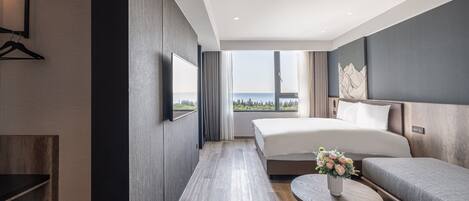 A modern hotel room with a large bed, a sofa, a small table with a vase, and a view of the sea.