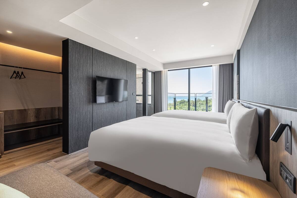 Quarto de hotel moderno com uma cama grande, televisão de tela plana, varanda com vista e piso de madeira.