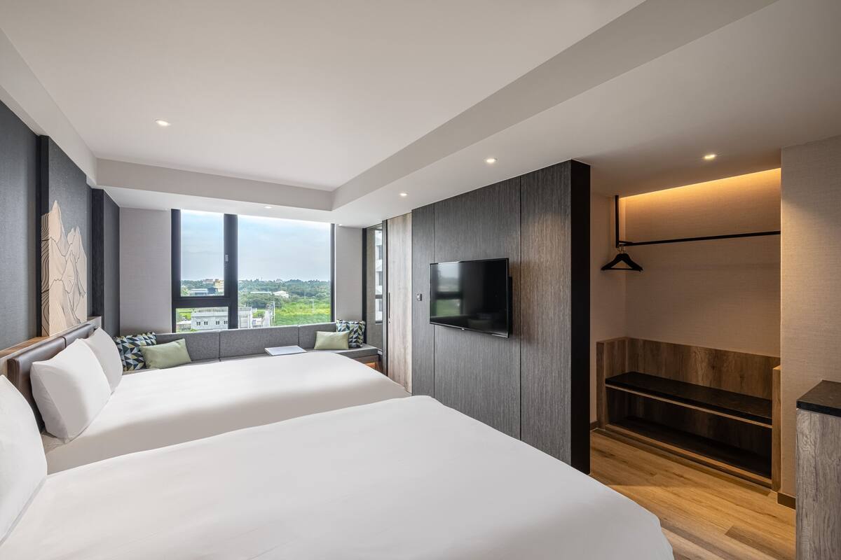 Quarto de hotel moderno com duas camas, uma janela grande com vista para a cidade, uma televisão de tela plana fixada na parede e um banco de madeira.
