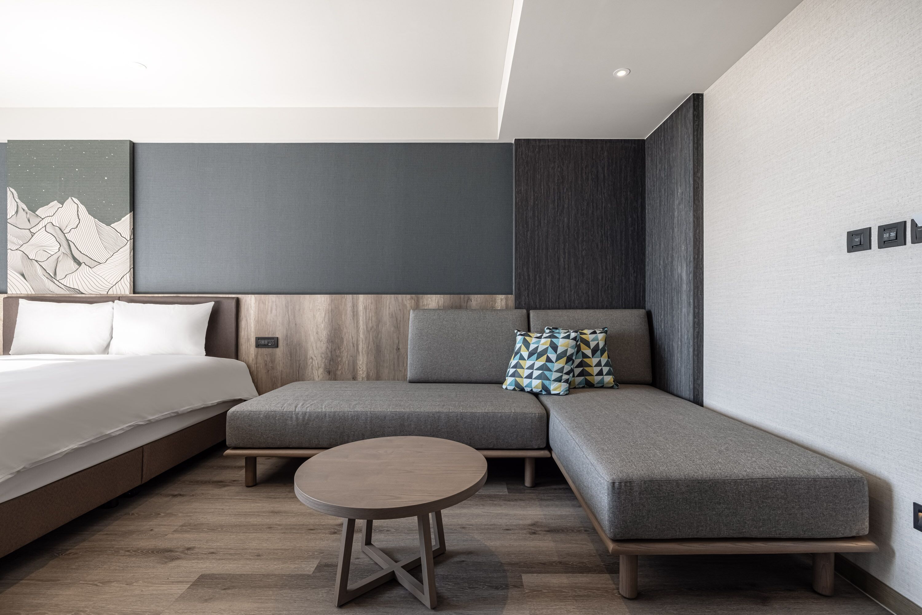 Ett modernt hotellrum med en säng, en grå soffa, ett litet runt bord och ett konstverk som hänger på väggen.