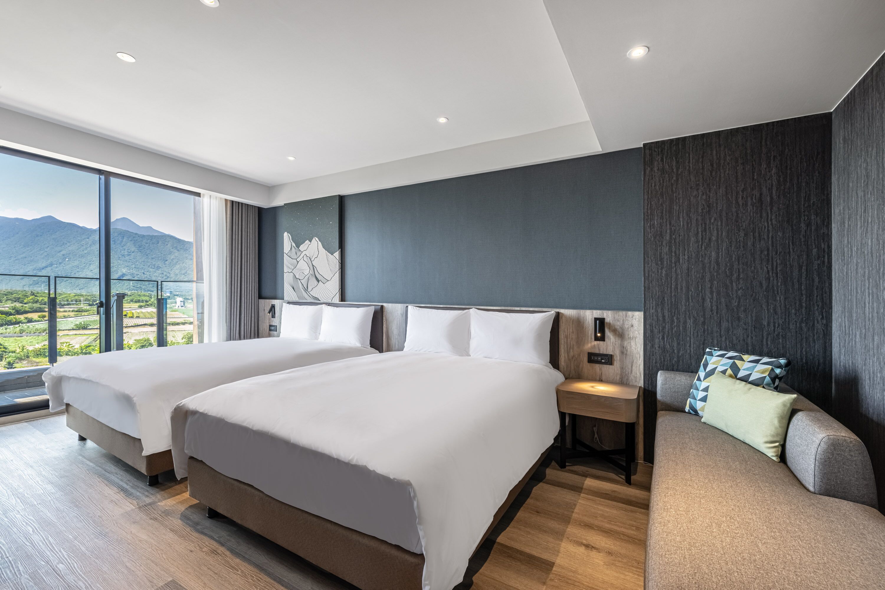 A modern hotel room with two beds, a sofa, and a large window offering a mountain view.